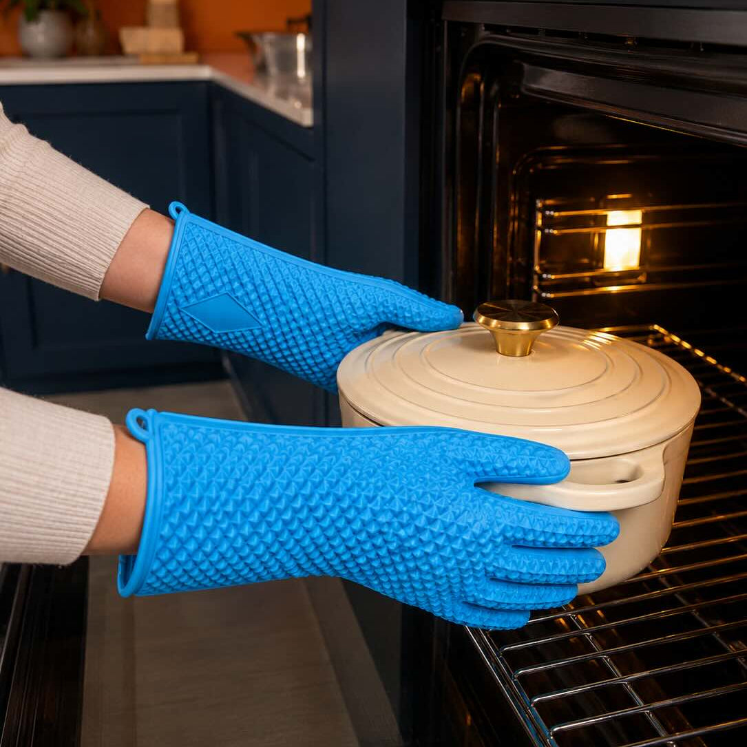 A person wearing bright blue silicone pot holders holds a cream-colored enameled cast iron Dutch oven with a lid while placing it into an open black oven. The background shows a modern kitchen setting with dark blue cabinets and a warm orange wall.