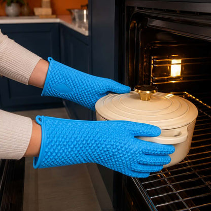 A person wearing bright blue silicone pot holders holds a cream-colored enameled cast iron Dutch oven with a lid while placing it into an open black oven. The background shows a modern kitchen setting with dark blue cabinets and a warm orange wall.