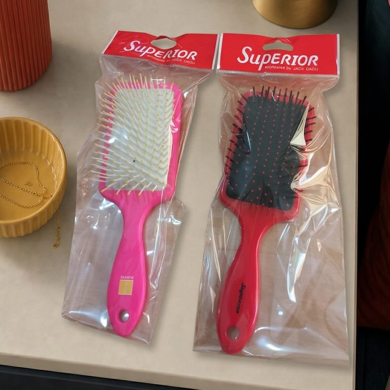 Two hairbrushes in packaging on a surface with a lamp and plant in the background.