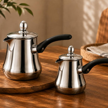 Two stainless steel Turkish coffee pots with black handles, one larger and one smaller, displayed on a wooden serving board on a wooden table with a warm, cozy background.