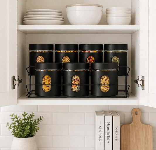 Black storage containers with food on a kitchen shelf with bowls and books in the background.