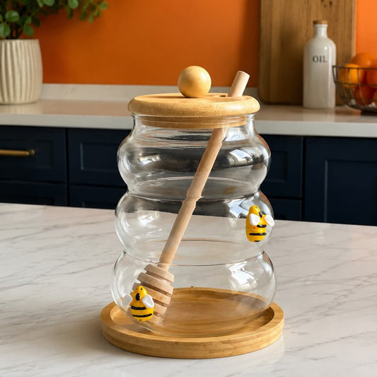 A clear glass honey jar with a wooden lid and base, a wooden dipper, and small yellow-and-black bee decorations, set on a white marble countertop with an orange wall and kitchen cabinets in the background.