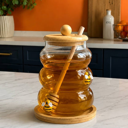 A clear glass honey jar with a wooden lid and dipper, filled with golden honey, set on a marble countertop in a kitchen with an orange wall and dark blue cabinets.