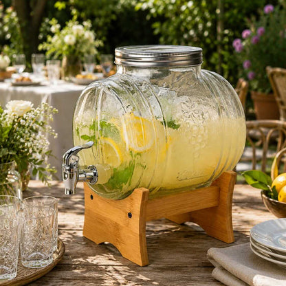 A large ribbed clear glass beverage dispenser filled with a pale yellow iced drink, set on a wooden stand outdoors on a table with plates, glasses, and greenery in the background.