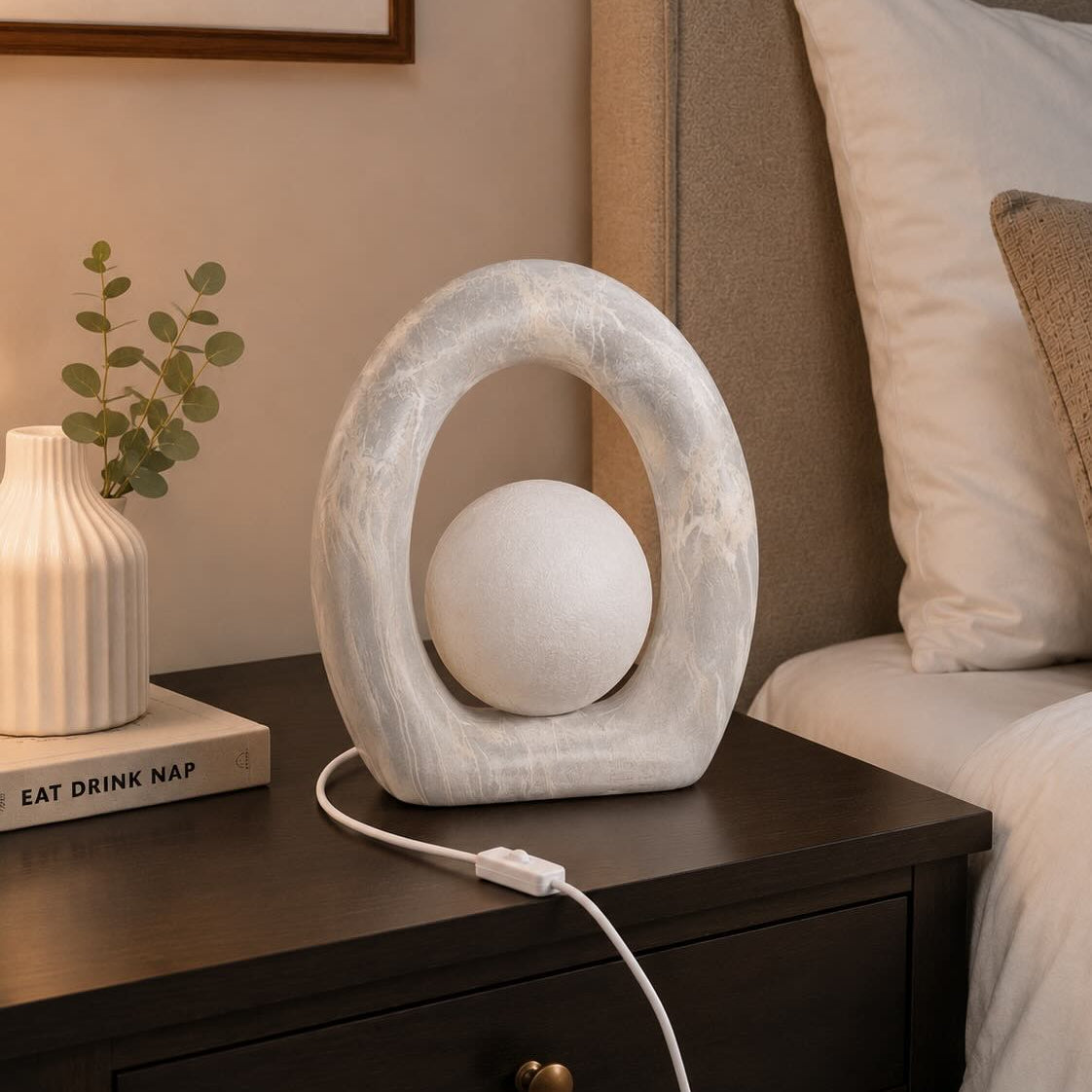 A modern oval-shaped white table lamp with a textured stone-like frame and a spherical shade, shown plugged into a dark wood bedside table in a bedroom setting.