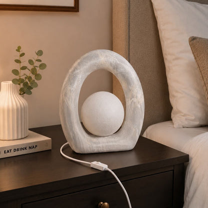 A modern oval-shaped white table lamp with a textured stone-like frame and a spherical shade, shown plugged into a dark wood bedside table in a bedroom setting.