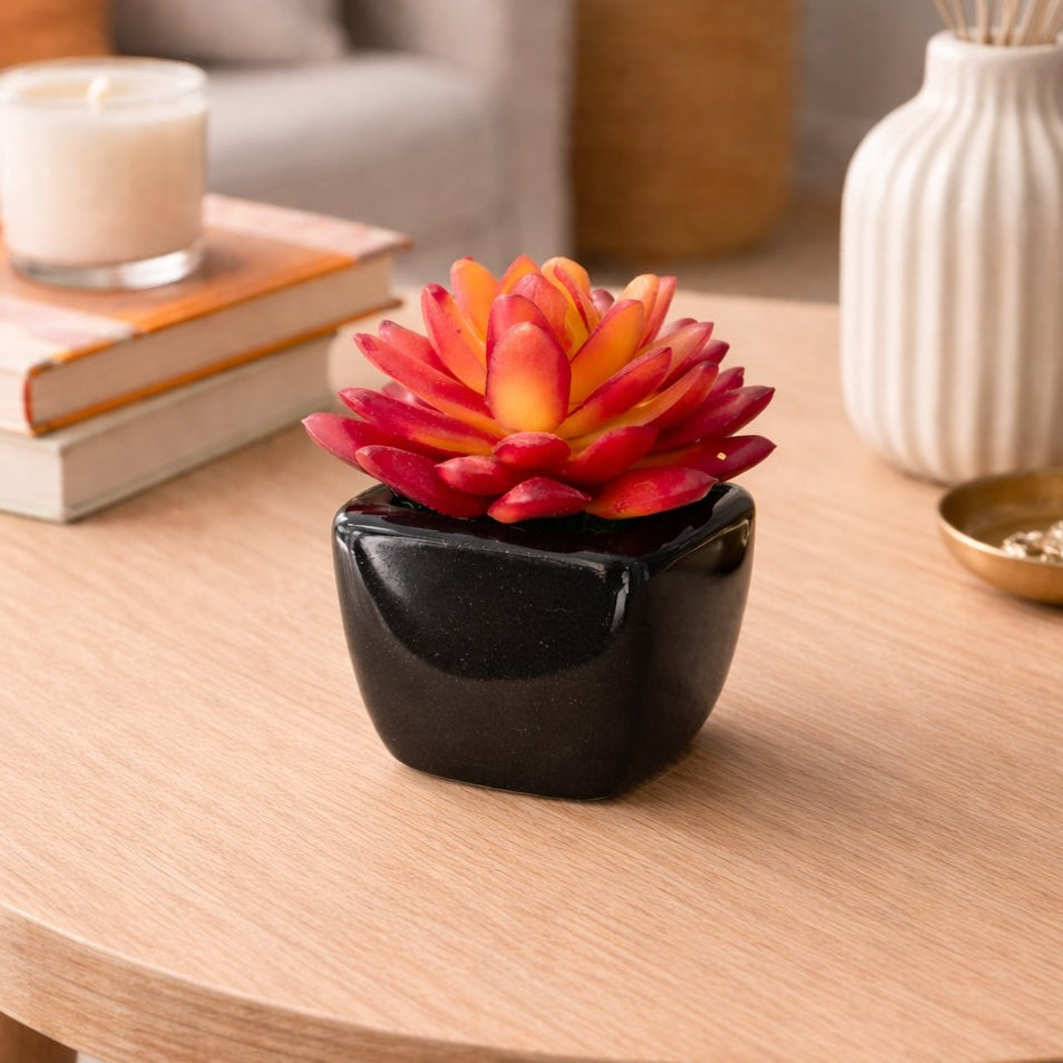 Decorative red and orange flower in a black pot on a wooden table with books and a candle in the background.