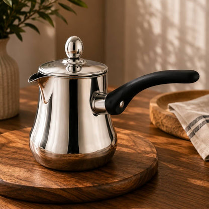 A polished stainless steel teapot with a black handle and lid knob, resting on a round wooden serving board on a warm wooden table.