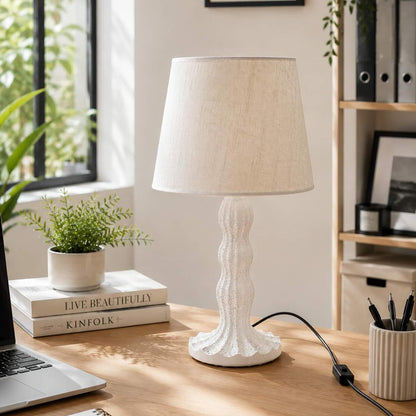 A white table lamp with a textured, sculptural base and a beige fabric drum shade sits on a wooden desk in a bright, modern home office setting.